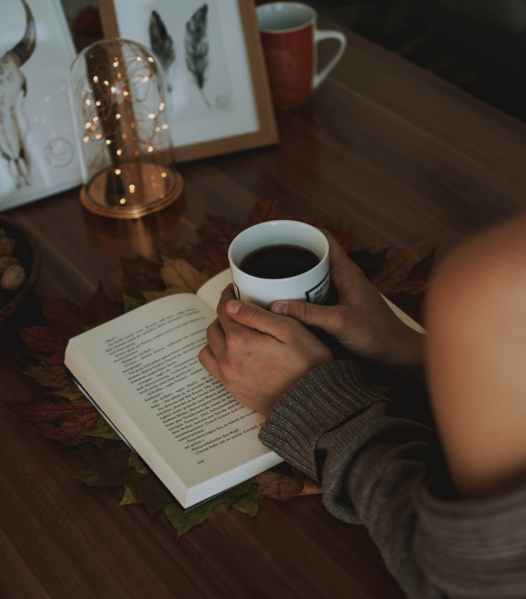girl holding mug of coffee above opened book on brown wooden table